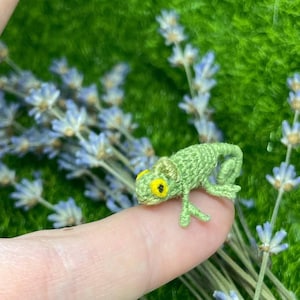 May include: A small green crocheted chameleon with yellow eyes sits on a finger. The chameleon is a miniature version of a real chameleon and is a handmade craft.
