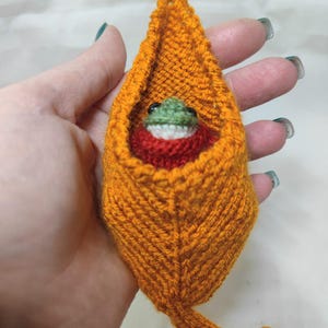 May include: A small, hand-knitted orange leaf-shaped pouch featuring a textured, ribbed pattern. Inside the pouch sits a tiny, round, green-and-white frog figure with black eyes, wearing a dark red collar or scarf around its neck.