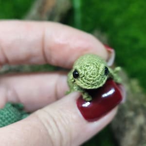 Op de afbeelding: Een kleine groene gehaakte kikker met zwarte ogen. De kikker wordt vastgehouden door een hand met rode nagellak.