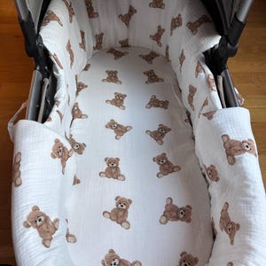 May include: A baby stroller interior with a white fabric lining. The fabric is patterned with numerous brown teddy bear illustrations. The stroller frame is gray, and the overall design is soft and inviting.