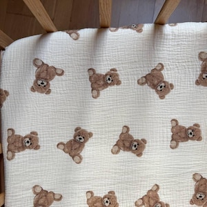 Puede incluir: Una sábana blanca para cuna con un patrón repetido de osos de peluche marrones. Los osos están en varias poses, creando un diseño lúdico y acogedor. La sábana está hecha de una tela suave y texturizada.