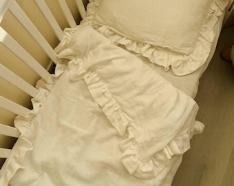 May include: Cream-colored baby bedding set with ruffled edges, including a pillow and a blanket, inside a white crib. The pillow has embroidered text. The bedding is made of a soft, textured fabric.