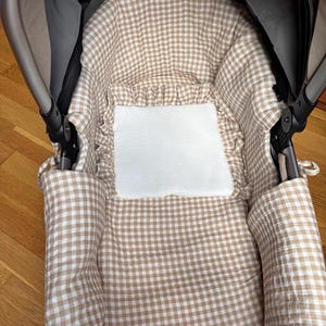 Op de afbeelding: De binnenkant van een kinderwagenbak met een beige en witte gingham ruitjesvoering en een ruche-detail. Een effen wit rechthoekig stoffen inzetstuk is in het midden van de gewatteerde bodem geplaatst, omringd door de stof met het ruitjespatroon.