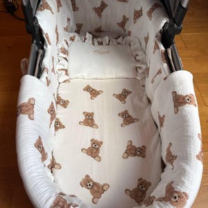 Puede incluir: Interior de cochecito de bebé con forro de tela blanca con un estampado repetido de ositos de peluche marrones. Se ve una pequeña almohada con el nombre "Capri" bordado. El cochecito tiene un diseño suave y acolchado.