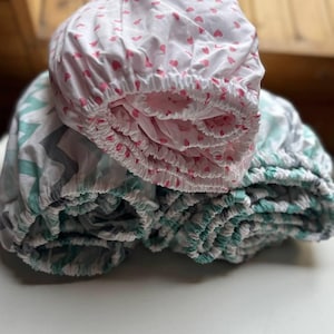 Może przedstawiać: Dwa prześcieradła z gumką, jedno z wzorem różowych serduszek, a drugie z zielono-szarym wzorem geometrycznym.