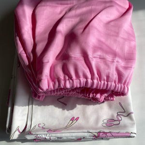 May include: A pink fitted sheet with a white sheet with a ballerina print underneath. The ballerina print features pink ballerinas with pink ribbons.