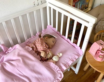 May include: A white crib with a pink ruffled bedding set. A young child is sleeping in the crib with a stuffed bunny toy. The crib is in a nursery with a bookshelf and a wooden toy kitchen.
