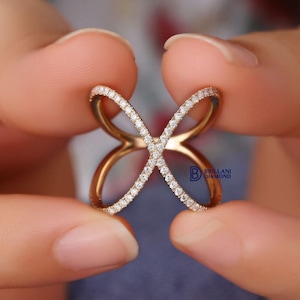 Puede incluir: Un anillo dorado con dise&ntilde;o de mariposa, sostenido entre dos manos. El anillo presenta un dise&ntilde;o entrecruzado con una fila de peque&ntilde;as piedras transparentes a lo largo de los bordes de las alas de la mariposa.