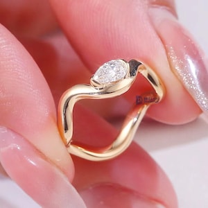 May include: A gold ring with a pear-shaped diamond. The ring has a unique, flowing design. The diamond is set in a bezel setting, and the band has a wavy shape.