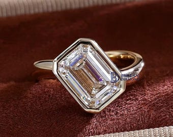 May include: A gold ring featuring a large, rectangular emerald-cut diamond set in a smooth, polished bezel setting. The center stone is clear and faceted, reflecting light with sharp, linear patterns. The band is a solid, rounded gold metal.