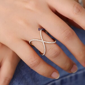 Puede incluir: Un anillo dorado con un dise&ntilde;o en forma de cruz, incrustado con peque&ntilde;as y brillantes piedras blancas. El anillo se lleva en un dedo, mostrando su estilo elegante y moderno. Las piedras preciosas capturan la luz, creando un efecto deslumbrante.