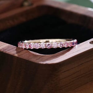 May include: A gold ring featuring a continuous row of pink gemstones. The ring is presented in a wooden jewellery box, highlighting its refined design. The gemstones are uniformly set, forming a seamless band of colour, adding to its elegance.