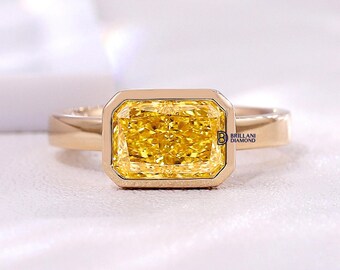 May include: A gold ring featuring a rectangular yellow gemstone set in a bezel setting. The stone has a faceted cut and a vibrant yellow hue. The band is smooth and polished gold. The text BRILLANI DIAMOND is printed on the side of the setting.