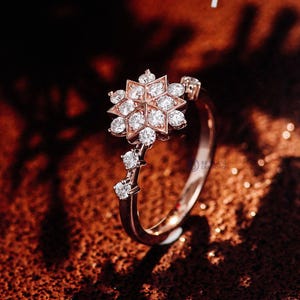 May include: A rose gold ring featuring a central snowflake-shaped cluster of round brilliant-cut diamonds. The band includes two additional small round diamonds set along one side, leading up to the main setting, with a polished metal finish.