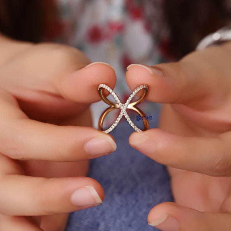 Puede incluir: Un anillo dorado con dise&ntilde;o de mariposa, sostenido entre dos manos. El anillo presenta un dise&ntilde;o entrecruzado con una fila de peque&ntilde;as piedras transparentes a lo largo de los bordes de las alas de la mariposa.