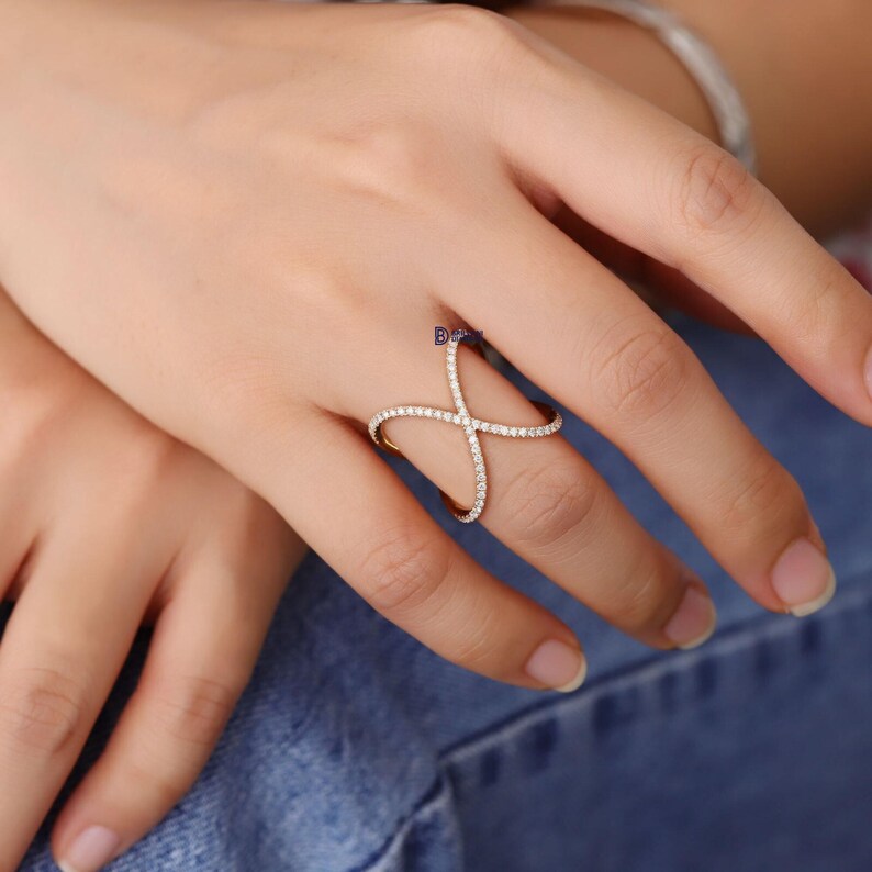 Puede incluir: Un anillo dorado con un dise&ntilde;o en forma de cruz, incrustado con peque&ntilde;as y brillantes piedras blancas. El anillo se lleva en un dedo, mostrando su estilo elegante y moderno. Las piedras preciosas capturan la luz, creando un efecto deslumbrante.