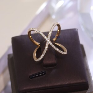 Puede incluir: Un anillo dorado con un dise&ntilde;o calado &uacute;nico. El anillo presenta una serie de l&iacute;neas curvas e intersecantes, incrustadas con peque&ntilde;as y brillantes gemas. Se exhibe sobre un soporte marr&oacute;n, resaltando su estilo elegante y moderno.