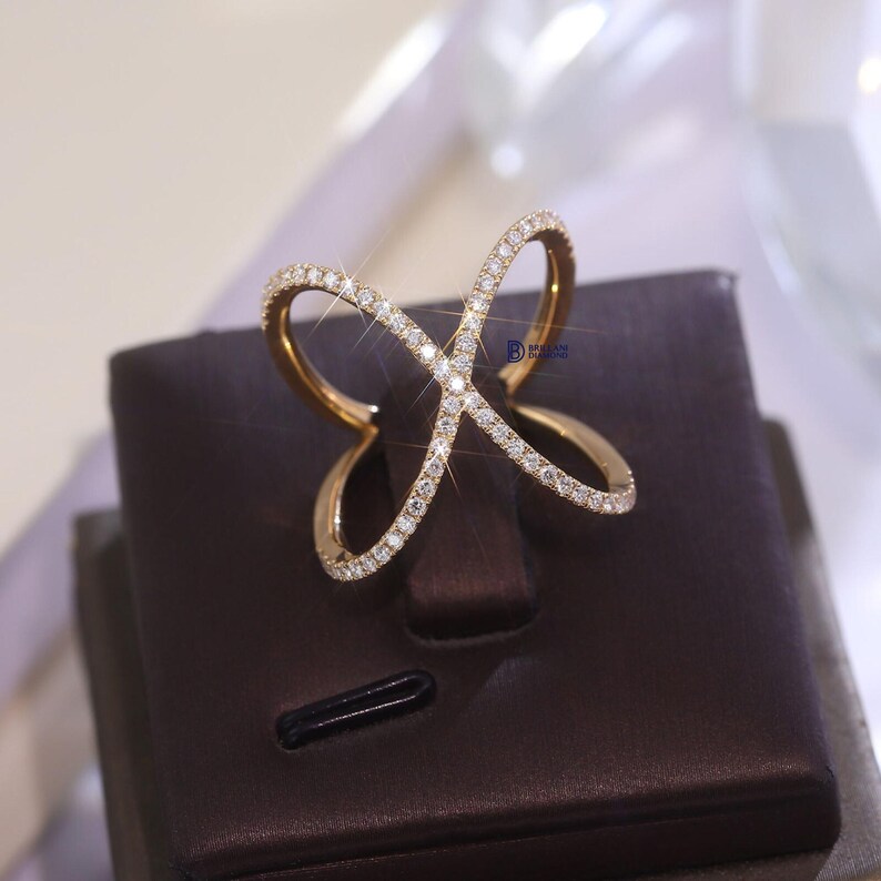 Puede incluir: Un anillo dorado con un dise&ntilde;o calado &uacute;nico. El anillo presenta una serie de l&iacute;neas curvas e intersecantes, incrustadas con peque&ntilde;as y brillantes gemas. Se exhibe sobre un soporte marr&oacute;n, resaltando su estilo elegante y moderno.