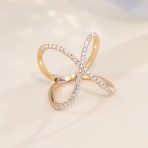 Puede incluir: Un anillo dorado con un dise&ntilde;o &uacute;nico e entrelazado. El anillo est&aacute; incrustado con peque&ntilde;as y brillantes piedras blancas. El dise&ntilde;o del anillo forma una forma abstracta, creando un aspecto moderno y elegante. El anillo est&aacute; sobre un fondo suave y neutro.
