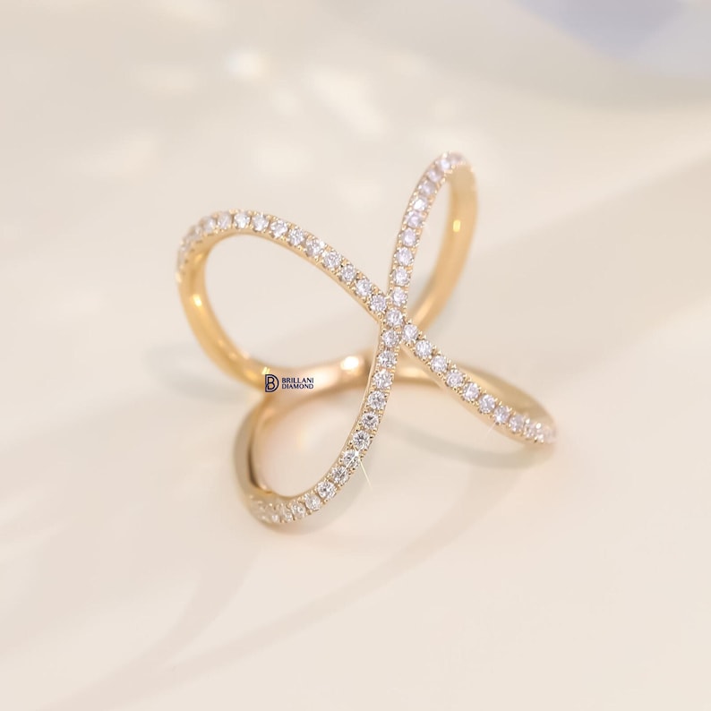 Puede incluir: Un anillo dorado con un dise&ntilde;o &uacute;nico e entrelazado. El anillo est&aacute; incrustado con peque&ntilde;as y brillantes piedras blancas. El dise&ntilde;o del anillo forma una forma abstracta, creando un aspecto moderno y elegante. El anillo est&aacute; sobre un fondo suave y neutro.