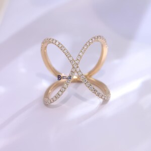 Puede incluir: Un anillo dorado con un dise&ntilde;o &uacute;nico en forma de cruz. El anillo est&aacute; adornado con numerosos diamantes peque&ntilde;os y brillantes, creando una exhibici&oacute;n brillante. El dise&ntilde;o del anillo presenta dos bucles superpuestos, formando una forma de X.