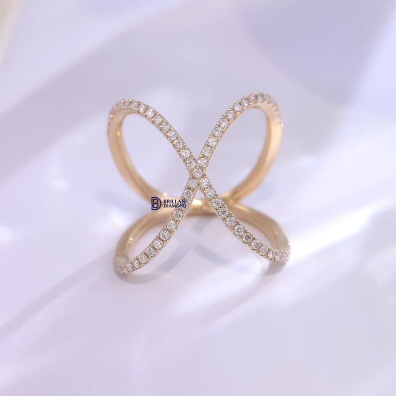 Puede incluir: Un anillo dorado con un dise&ntilde;o &uacute;nico en forma de cruz. El anillo est&aacute; adornado con numerosos diamantes peque&ntilde;os y brillantes, creando una exhibici&oacute;n brillante. El dise&ntilde;o del anillo presenta dos bucles superpuestos, formando una forma de X.