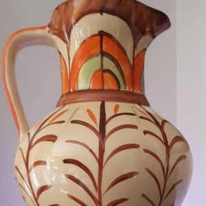 Peut inclure: Un pichet en céramique de couleur crème avec une anse brune et un motif géométrique dans les tons orange, vert et marron. Le pichet présente un motif de feuilles stylisé sur le corps.