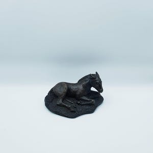 May include: A dark brown sculpture of a resting foal. The foal is lying down on a textured base. The sculpture has detailed features and is set against a white background.