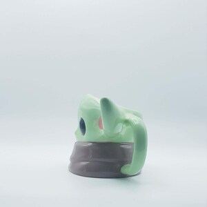 May include: A ceramic mug shaped like a character with light green skin, large dark eyes, and pointed ears. The mug features a smooth, glossy finish and a wide, rounded handle on the side. The base of the mug is a solid, dark gray color.