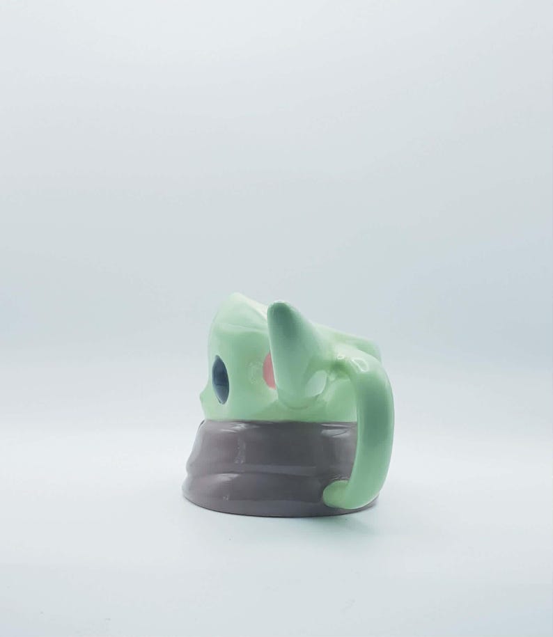 May include: A ceramic mug shaped like a character with light green skin, large dark eyes, and pointed ears. The mug features a smooth, glossy finish and a wide, rounded handle on the side. The base of the mug is a solid, dark gray color.