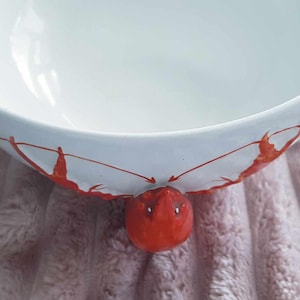 May include: White ceramic bowl with a red lobster design. The bowl is empty and sitting on a white fluffy surface.