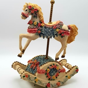 May include: A cream-coloured carousel horse figurine mounted on a central gold pole, featuring a blue saddle blanket adorned with pink, yellow, and purple roses. The horse is attached to a rocking base decorated with floral garlands and ribbons.
