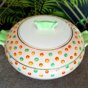 Könnte beinhalten: Eine weiße Keramik-Servierplatte mit einem grünen Deckel und einem Muster aus orangefarbenen, gelben und grünen Punkten auf einem weißen Gitter. Die Platte hat einen goldenen Rand.