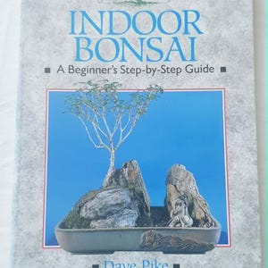 May include: A softcover book titled Indoor Bonsai: A Beginner's Step-by-Step Guide by Dave Pike. The cover features a light gray textured background and a central image of a bonsai tree planted in a rectangular pot with rocks and a small figurine.