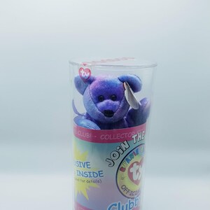 May include: A purple plush bear toy with a black nose and ears is inside a clear plastic container. The container has a label with the text "Join the Beanie Club" and "Exclusive Inside". A heart-shaped logo is visible on the container.
