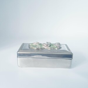 Peut inclure: Une boîte rectangulaire en métal argenté avec une finition lisse et polie. Le couvercle présente un ornement en relief au centre, représentant des motifs floraux et des feuilles détaillés peints dans des tons rose pâle et vert sauge.