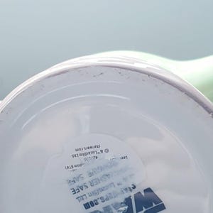 May include: The underside of a white ceramic mug featuring the blue Star Wars logo. A clear sticker is applied to the base with text that reads Microwave Safe, Dishwasher Safe, and includes copyright information for Lucasfilm Ltd. and starwars.com.