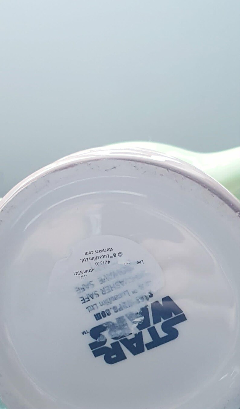 May include: The underside of a white ceramic mug featuring the blue Star Wars logo. A clear sticker is applied to the base with text that reads Microwave Safe, Dishwasher Safe, and includes copyright information for Lucasfilm Ltd. and starwars.com.