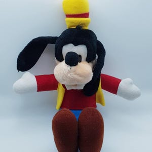 May include: A Goofy plush toy, a Disney cartoon character. The toy features a black head and ears, a yellow hat, and a red shirt with a yellow vest. The arms are red with white hands, and the legs are brown with blue shorts.