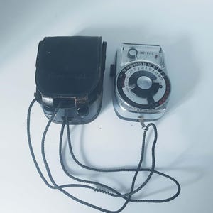 May include: A silver Imperial light meter with a circular dial featuring black and red numerical markings, a central rotating knob, and a black neck strap. Beside it sits a black leather protective case with a matching black cord strap.