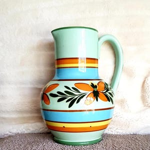 May include: A hand-painted ceramic jug with a light green base and handle. The jug features horizontal stripes in orange, yellow, and blue, and is decorated with orange floral designs and green leaves. The jug is approximately 25 cm tall.