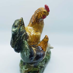 May include: A ceramic rooster figurine with a red comb and wattle, brown and tan body, and green base. The rooster is perched on a green and brown textured base, with a detailed design.