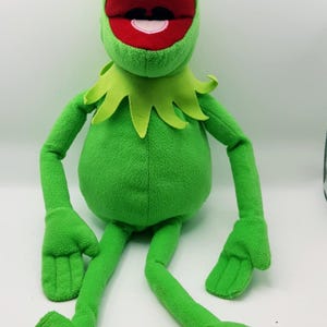 May include: A plush Kermit the Frog doll, featuring a bright green body, large red mouth, and white teeth. The doll has black eyes, a green collar, and long arms and legs with webbed feet. The toy is designed to resemble the Muppet character.