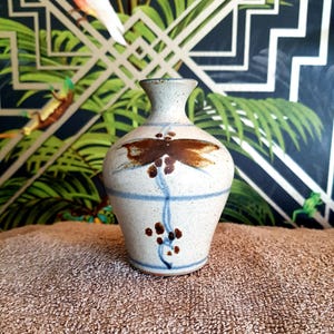 May include: A speckled ceramic vase with a brown dragonfly design and blue accents. The vase has a rounded body and a narrow neck. The dragonfly is painted on the front of the vase, with blue lines and brown dots below.