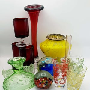 May include: A collection of glassware including a tall red vase, a yellow flower frog bowl, a green glass candle holder with butterfly motifs, two glass spheres with internal patterns, and several clear drinking glasses with red and yellow floral prints.