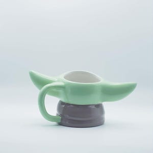 May include: A ceramic mug shaped like a character with large pointed ears, featuring a light green upper section and a smooth dark gray base. The mug includes a curved handle on the side and a white interior, designed with a wide opening at the top.