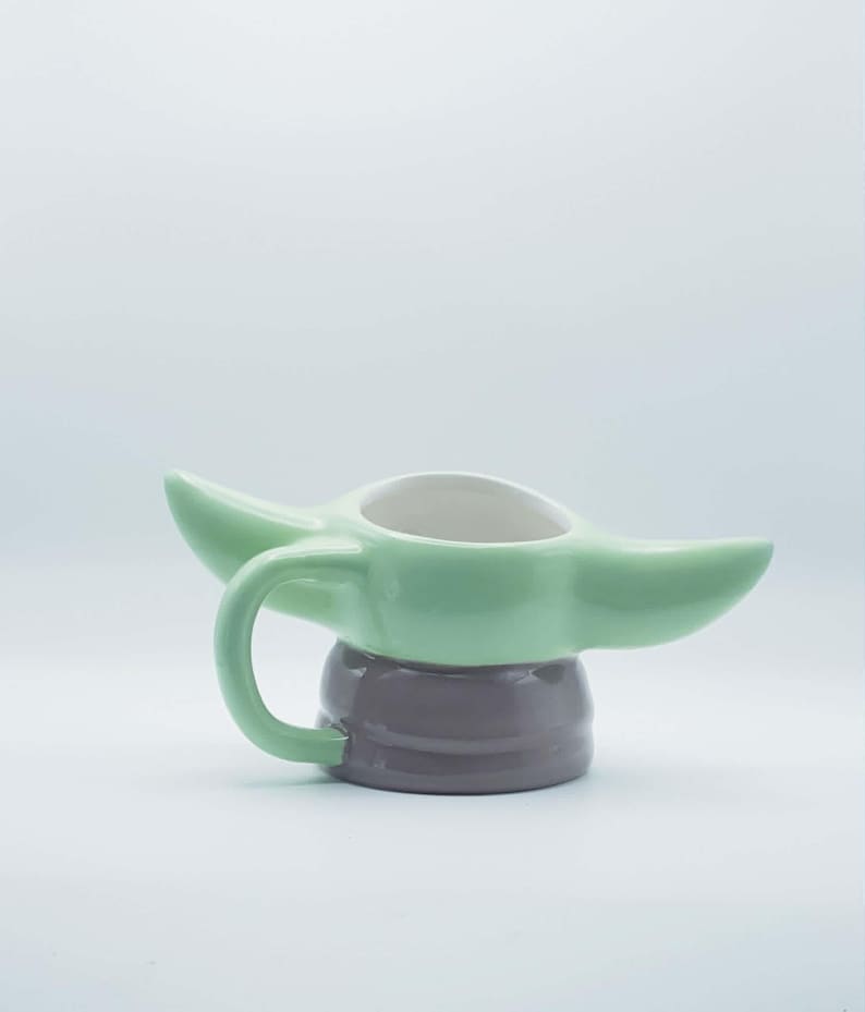 May include: A ceramic mug shaped like a character with large pointed ears, featuring a light green upper section and a smooth dark gray base. The mug includes a curved handle on the side and a white interior, designed with a wide opening at the top.