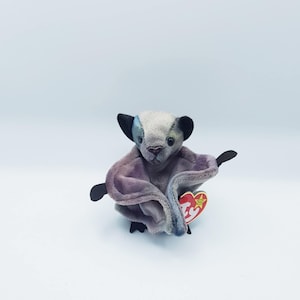 Könnte beinhalten: Ein Plüschtier in Form einer Fledermaus mit grauem Kopf, lila Körper und schwarzen Flügeln. Die Fledermaus hat ein rotes herzförmiges Etikett.