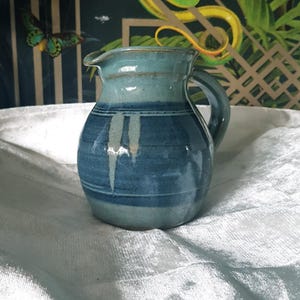 May include: A handcrafted ceramic jug with a rounded form and a handle. The jug is glazed in shades of blue, with darker blue horizontal stripes around the middle. The spout is slightly flared. A decorative piece for the kitchen or dining room.