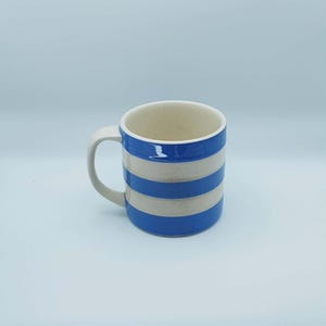 May include: A ceramic mug with a white handle and interior. The exterior features alternating horizontal stripes of blue and off-white. The mug is designed for hot beverages and has a simple, classic design.